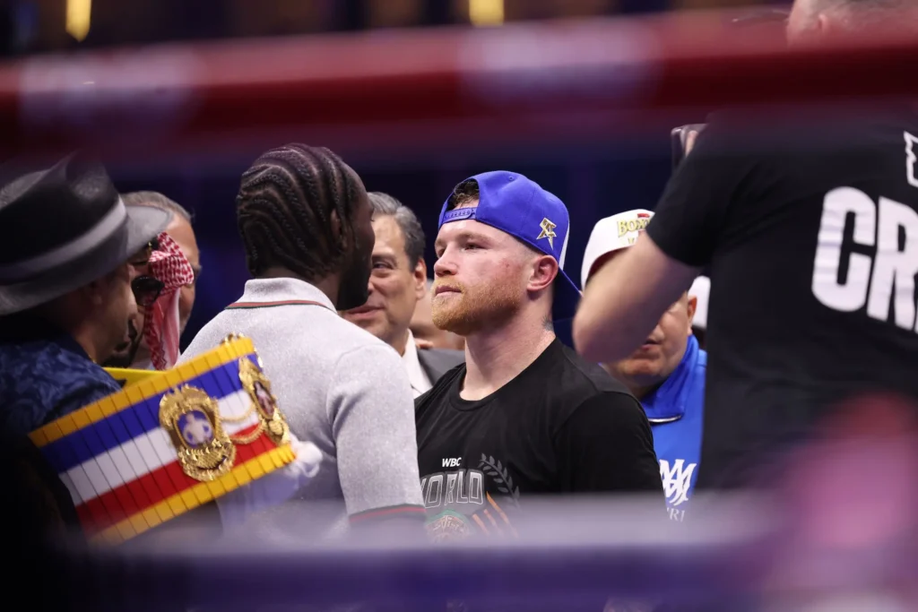 Canelo Álvarez, in a blue cap, stares down Terence Crawford in the ring as entourages and officials look on.