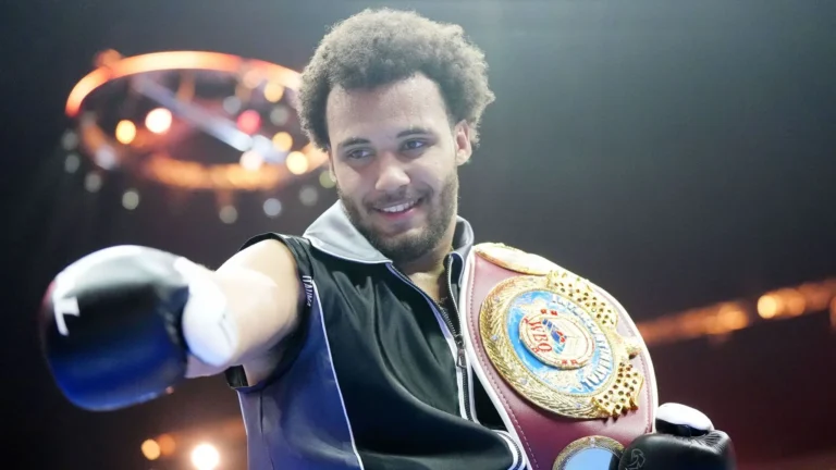 Moses Itauma smiles while wearing a championship belt over his shoulder and pointing toward the camera, dressed in black boxing gear inside a lit-up arena.